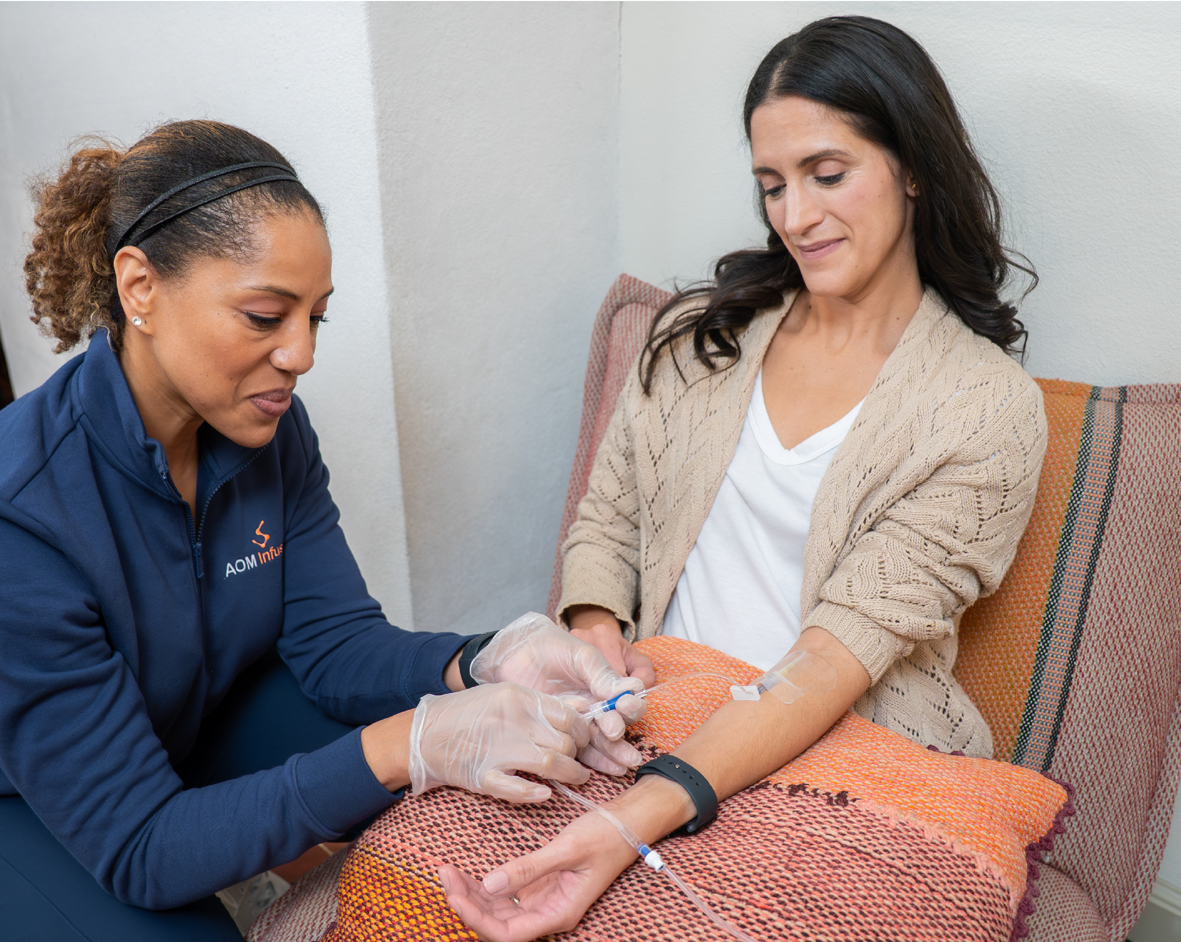 A nurse wearing gloves and an AOM Infusion jacket administers an IV infusion into a woman’s arm as the woman sits comfortably in a chair with her arm resting on an orange pillow.